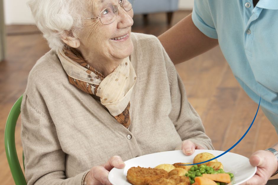 Seniorin hält einen Teller mit Essen in der Hand und wendet sich lächelnd einer Pflegerin zu - Leistung Verköstigung