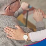 Nahaufnahme von hinten. Frau legt fürsorglich die Hand auf den Rücken eines alten Mannes zum Thema Rundumbetreuung24 Leistungen